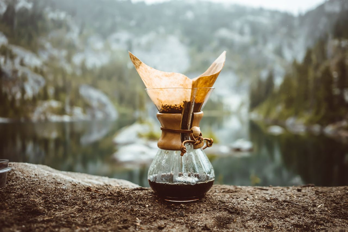 chemex coffee maker sitting on log in front of an alpine lake