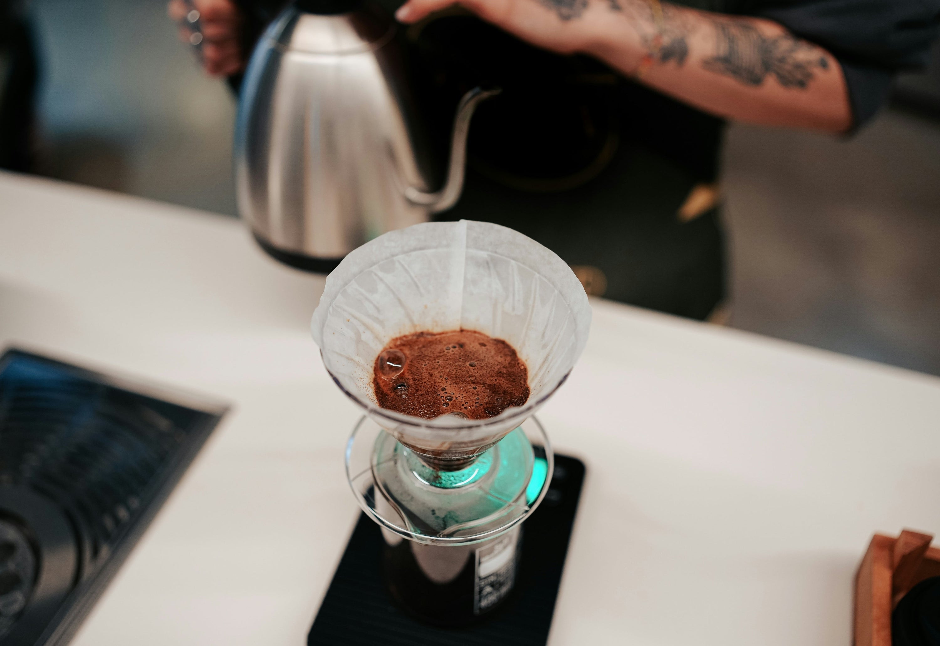 person holding gooseneck kettle beside pour over coffee being bloomed